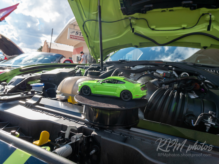 A small lime green toy Mustang car sits on a pedestal in the engine bay for a full sized Mustang which is the same color.